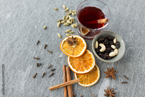 christmas and seasonal drinks concept - hot mulled wine, dry orange slices, raisins with cashew nuts and aromatic spices on grey background