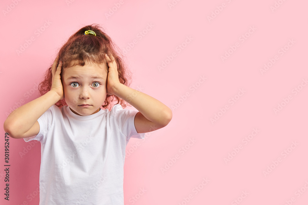 Frightened kid with auburn hair and big eyes, covered ears while standing on pink background Frightened kid with auburn hair and big eyes, covered ears while standing on pink background