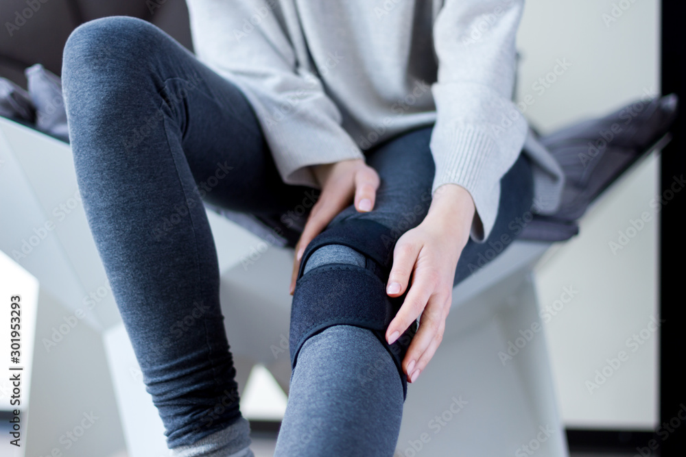 Closeup female leg in grey leggings dressed in knee brace to help promote  recovery of bones, muscles, ligaments. Woman is feeling pain in joints  after injury. Arthritis and meniscus diseases concept. Stock