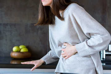 Young woman in grey clothes is holding hands on belly. Brunette girl is feeling bad and sick. Sudden onset of diarrhea, stomach ache, pancreatitis, appendicitis attack. Bad junk food concept.
