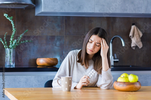 Brunette girl is sitting at table in kitchen and holding hand on head temple. Young woman is feeling bad and going to take painkiller pills. Sudden attack of migraine and headache. Effects of stress.