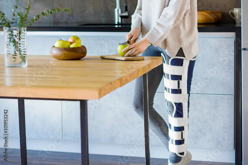 Girl with broken leg is standing in kitchen and cooking. Injured woman wearing supporting compression bandage for trauma to help promote recovery of bones, muscles, ligaments. Orthopedic diseases. photo
