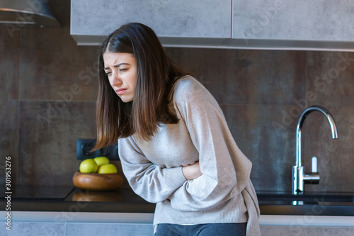 Young woman in grey clothes is holding hands on belly. Brunette girl is feeling bad and sick. Sudden onset of diarrhea, stomach ache, pancreatitis, appendicitis attack. Bad junk food concept. photo