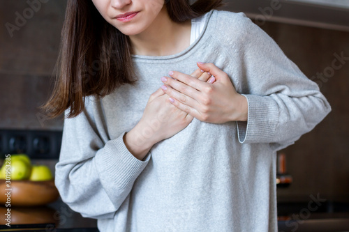 Young woman in grey clothes is holding hands on breast. Brunette girl is feeling bad. Sudden heart attack, myocardial infarction at home. Effect of stress and unhealthy lifestyle concept.