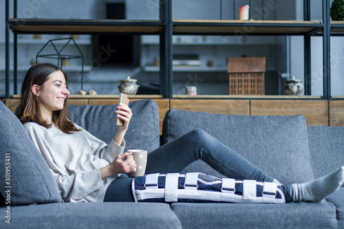 Papier peint Smiling brunette girl with broken leg is sitting on couch sofa, resting and drinking tea at home