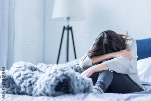 Young woman in gray clothes is sitting curled up on bed at home. Upset brunette girl is suffering, crying. Depression because of loneliness, stress, problems. Psychological disorders concept. photo
