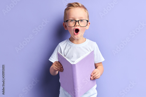 Fotografi Lovely boy wearing white t-shirt and glasses on eyes reading book with purple cover, stand in shock after reading, bright emotions of kid boy