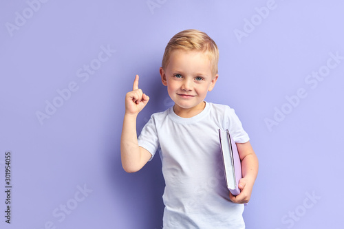 Caucasian kid show bright emotions standing over purple background, holding purple notebook.