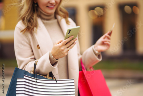 Hands of beautiful woman with telephone and a credit card. Young woman holding shopping bags on the city street. Online shopping concept. Consumerism, purchases, shopping, lifestyle concept.