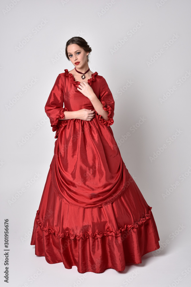 full length portrait of a brunette girl wearing a red silk victorian gown. Standing pose on a white studio background. full length portrait of a brunette girl wearing a red silk victorian gown. Standing pose on a white studio background.