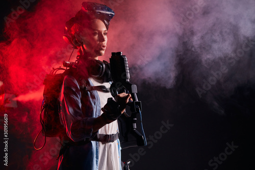 Virtual reality, hi-tech, innovation, future concept. Young caucasian woman holding weapon, look side, Stand isolated over red smoky background