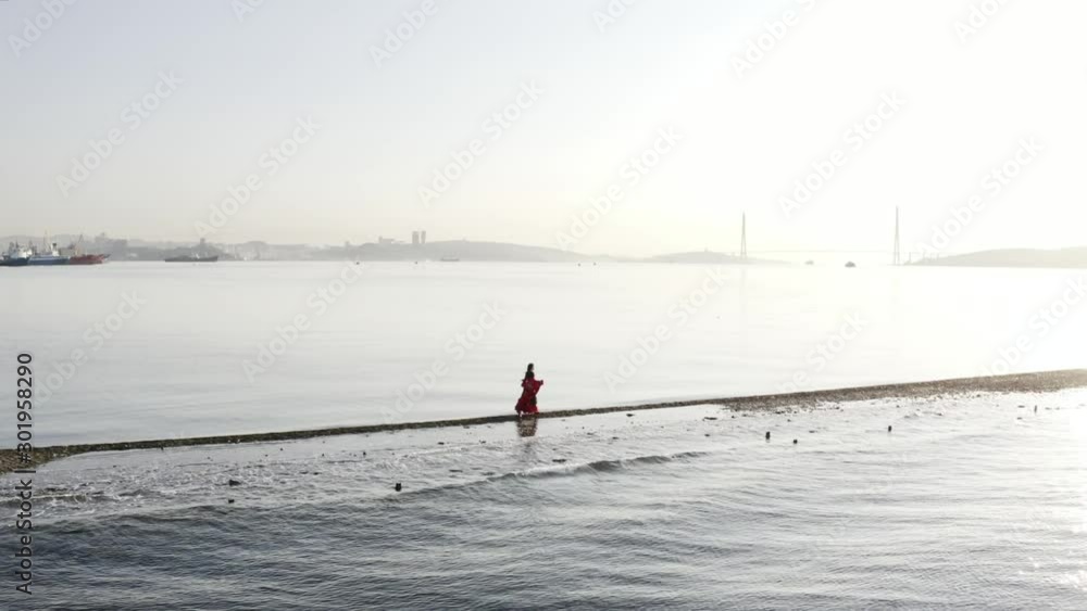 Aerial view of gipsy woman in bright dress running along the sandbar of lighthouse. Morning. Calm sea. Cityscape of Vladivostok is on the background. Aerial view of gipsy woman in bright dress running along the sandbar of lighthouse. Morning. Calm sea. Cityscape of Vladivostok is on the background.