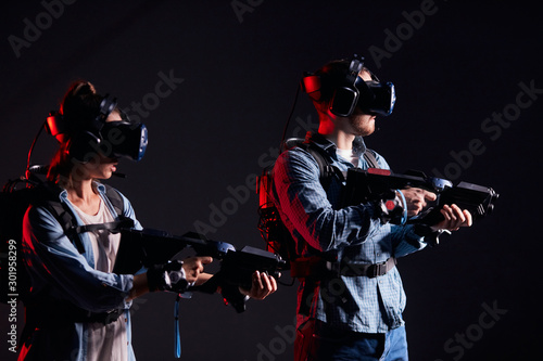 Couple of virtual reality gamers stand in neon backlit. Man and woman with virtual weapons isolated over black backgrond