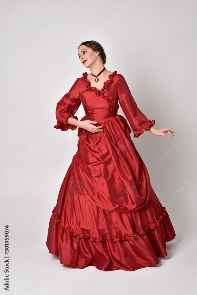 full length portrait of a brunette girl wearing a red silk victorian gown. Standing pose on a white studio background. full length portrait of a brunette girl wearing a red silk victorian gown. Standing pose on a white studio background.
