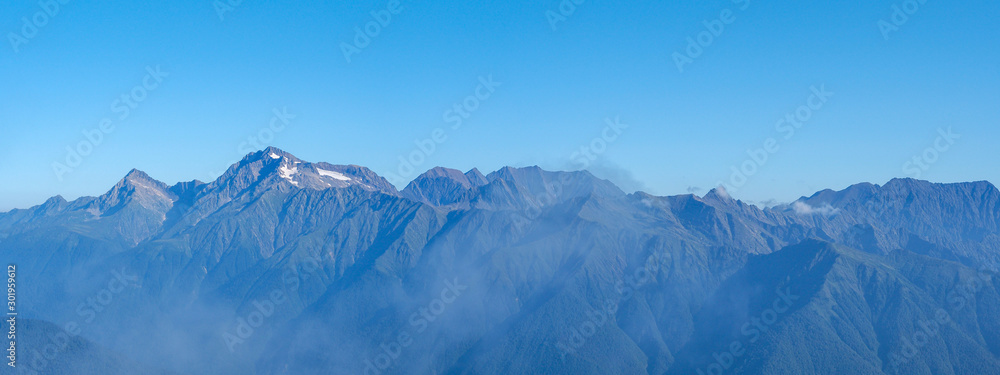 Fototapeta premium Panorama of mountain ranges in sunny early morning; clouds floating on peaks, first sunrise sunbeams; Chugush mount is highest point in area; spruce-fir forests on slopes; higher subalpine meadows