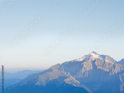Panorama of mountain ranges in sunny early morning; clouds floating on peaks, first sunrise sunbeams; Chugush mount is highest point in area; spruce-fir forests on slopes; higher subalpine meadows