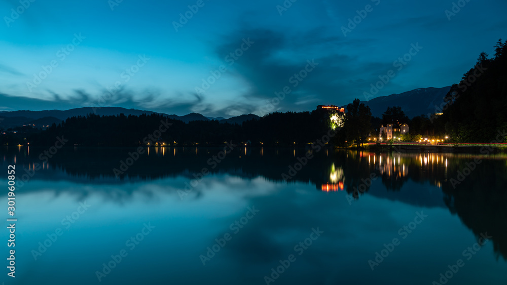 Bled lake landscape at dusk, in the blue hour. You can see the hills, Bled Castle, and some clouds; as well as its reflection in the water.