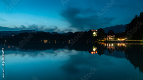 Bled lake landscape at dusk, in the blue hour. You can see the hills, Bled Castle, and some clouds; as well as its reflection in the water.