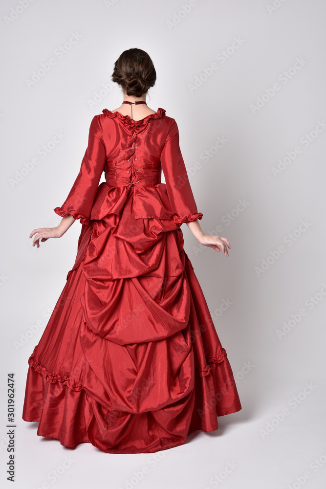 full length portrait of a brunette girl wearing a red silk victorian gown. Standing pose, with back to the camera,  on a white studio background.