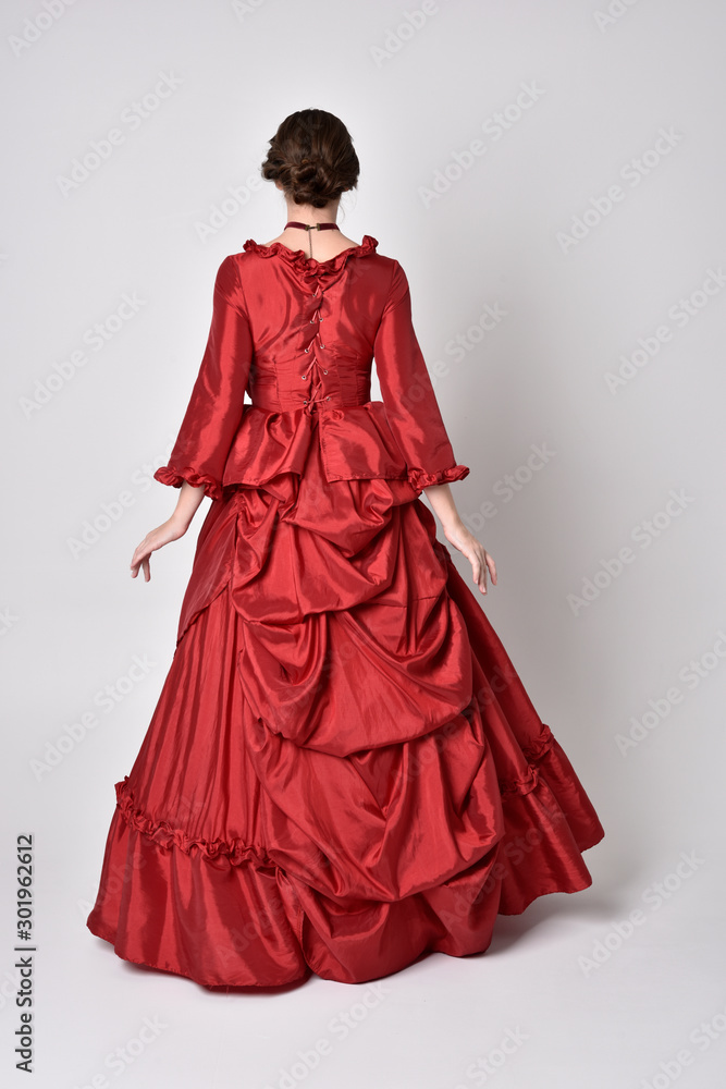 full length portrait of a brunette girl wearing a red silk victorian gown. Standing pose, with back to the camera, on a white studio background. full length portrait of a brunette girl wearing a red silk victorian gown. Standing pose, with back to the camera, on a white studio background.