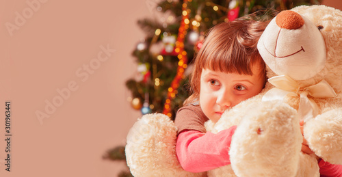 Christmas picture of little beautiful girl hugging teddy bear at home and waiting for Christmas and Santa Claus. Free space for text.