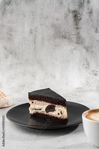 Obraz na płótnie Creamy cheesecake with chocolate cookies and cream biscuits, chocolate cookies and cream biscuits cheese cake. Piece of cake on black plate, white cup on white marble background. Vertical photo. w salonie