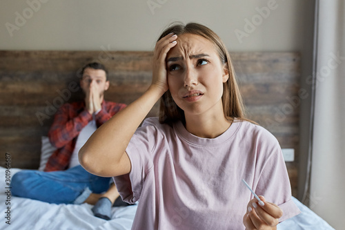 Young upset woman got positive result of pregnancy test, keeps hand on forehead with puzzled expression, demonstrating sadness, looking up with terrified feelings, close up,