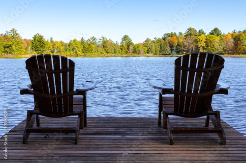 Fototapeta Naklejka Na Ścianę i Meble -  Two Muskoka chairs sitting on a wood dock facing a lake in a calm autumn season sunny day