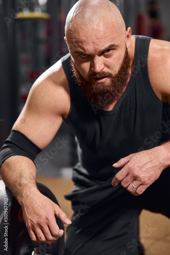 Brutal hairless bodybuilder with thick brunette beard looking straight at camera with evil powerful expression, ready to start competition in professional gym