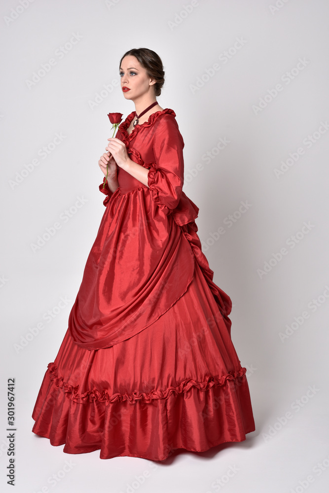 full length portrait of a brunette girl wearing a red silk victorian gown. holding a single rose on a white studio background. full length portrait of a brunette girl wearing a red silk victorian gown. holding a single rose on a white studio background.