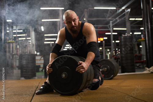 Young man with bald head, dressed in black stylish sportswear looks aside with interest, sitting on barbell, changing metal weight plate, gym atmosphere, indoor shot