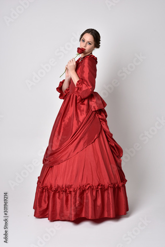 full length portrait of a brunette girl wearing a red silk victorian gown. holding a single rose on a white studio background.