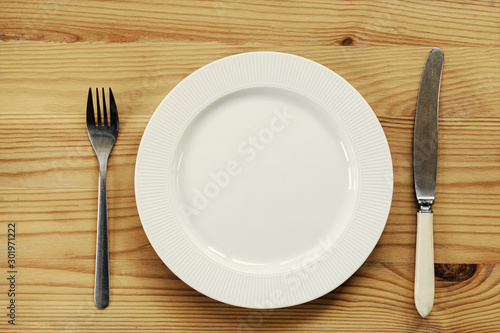 White plate and knife with a fork are served on a wooden table. view from above.