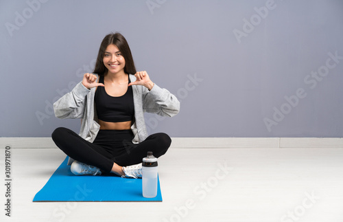 Young sport woman sitting on the floor with mat proud and self-satisfied