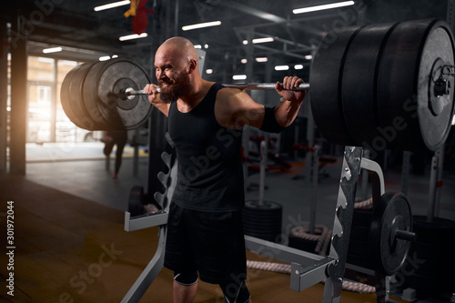 Hairless bald powerlifter with thick beard dressed in black sports clothes, attempting to rise heavy barbell, making effort, training hard, preparing for workout, side shot, professional sport concept
