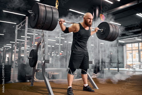 Brutal hairless young male athlete with thick beard raising hands up with heavy barbell, lifting barbell above head, looking away with strong face, expressing power, preparing for workout in gym