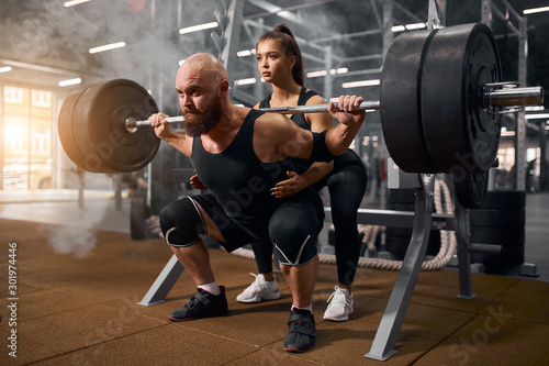 Strong brutal male powerlifter doing squats using heavy barbell, young active female trainer showing the right position, preparing for serious competition of weightlifting, professional sport