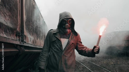 Man in gas mask walks through abandoned rusty carriages at destroyed train station red holding signal fire in hand. Armageddon, male survivor after nuclear or chemical disaster on deserted railway