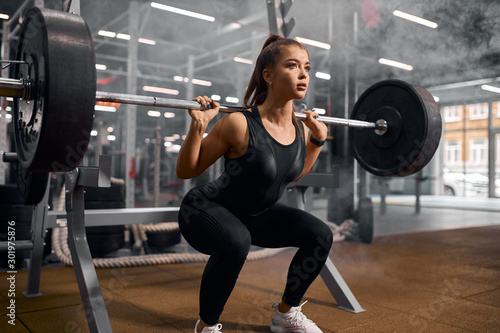 Fototapeta Naklejka Na Ścianę i Meble -  Charming strong female powerlifter dressed in black sportswear and white sneakers, doing squats, trying to stand with heavy barbell, professional sport concept, indoor shot