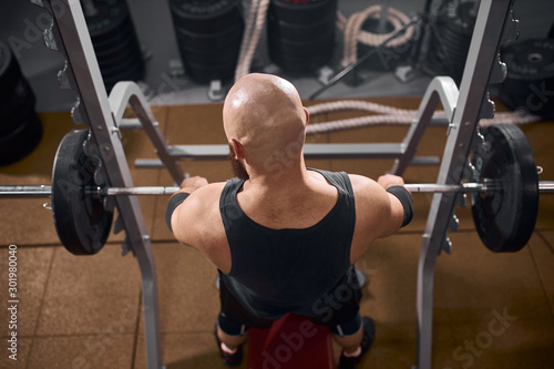 Back view of hairless bald man sitting in front of iron heavy barbell, keeping hands on metal bar, preparing to bench press, high angle view, indoor shot, fitness club concept