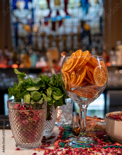 Glass full of dried sliced oranges  at a bar with rose petals sprinkled around for seasonal decoration. Bottles on shelves behind  out of focus.