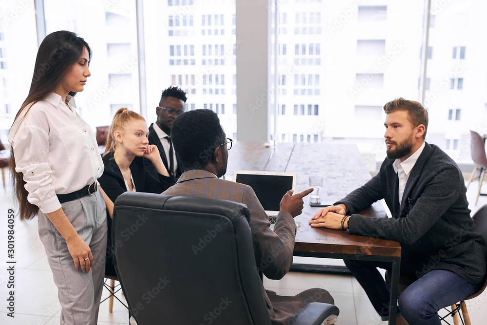 African male wearing tuxedo sit speaking with caucasian man while others listening to them. Teamwork in business center, window background African male wearing tuxedo sit speaking with caucasian man while others listening to them. Teamwork in business center, window background
