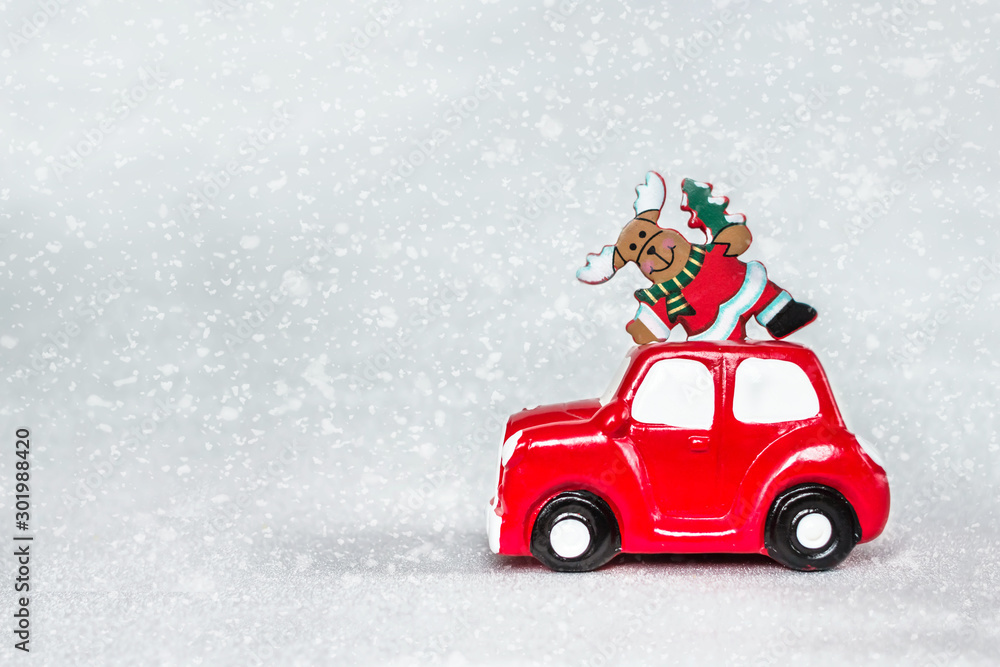 Obraz Red toy retro car delivering Christmas deer on the roof at snowy silver background. Merry Christmas and Happy New Year concept. Copy space.