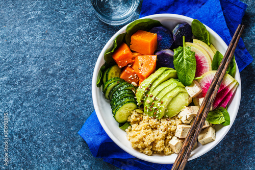 Quinoa salad with tofu, avocado and vegetables in white bowl, top view, blue background. Vegan breakfast.