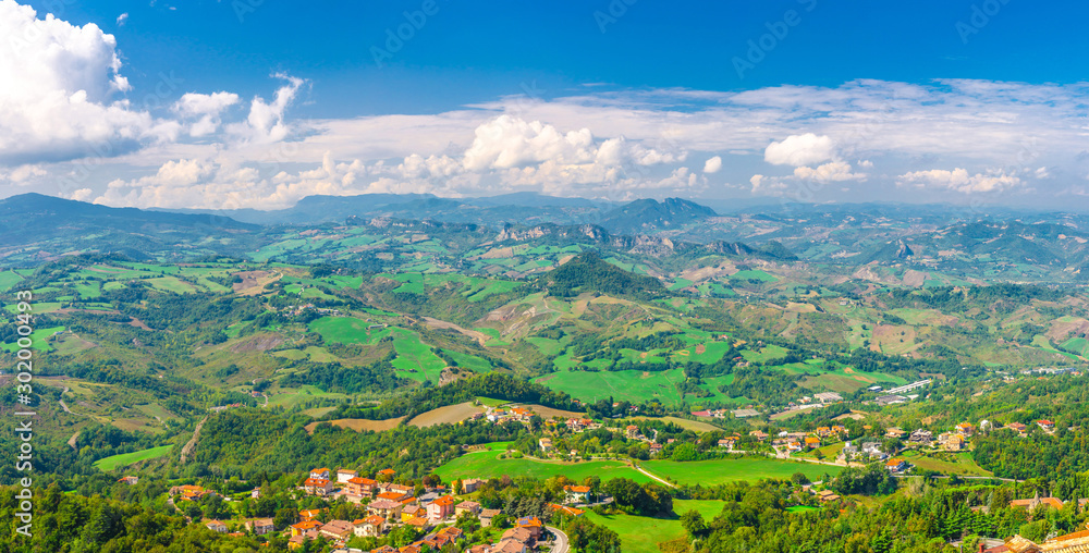 Aerial top panoramic view of landscape with valley, green hills, fields and villages of Republic San Marino suburban district with blue sky white clouds background. View from San Marino fortress.