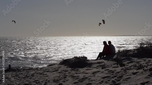 Human silhouettes, couple sitting on beach sand dune, grass blowing in wind. Kite surfer, kitesurfing in silver mirror glistening sea waters. Sun reflection on choppy ocean waves. African beach sunset
