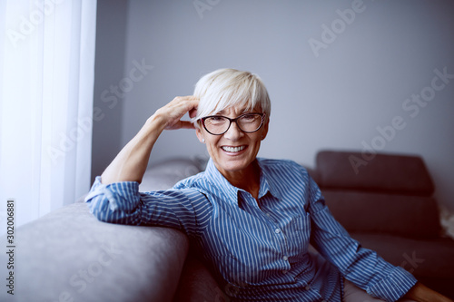 Portrait of smiling charming caucasian blond woman sitting on sofa in living room next to window and looking at camera. photo