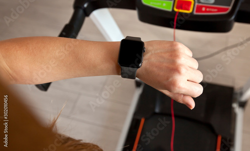The woman is running on treadmill at home and looking on her smartwatch on her hand