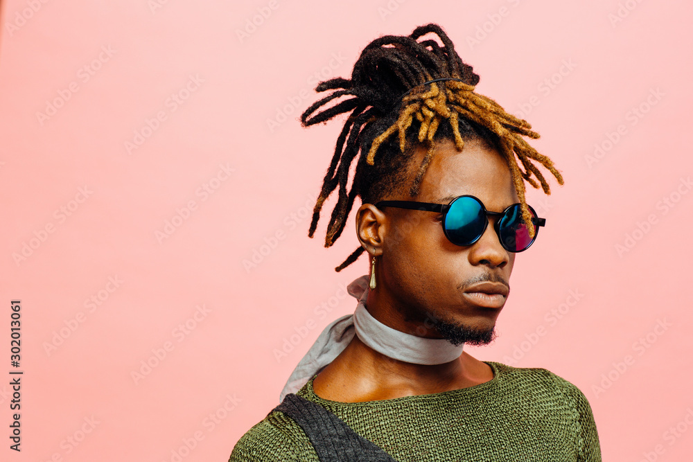 Close up portrait of a young man in green with dreadlocks and blue sunglasses, isolated on pink Close up portrait of a young man in green with dreadlocks and blue sunglasses, isolated on pink