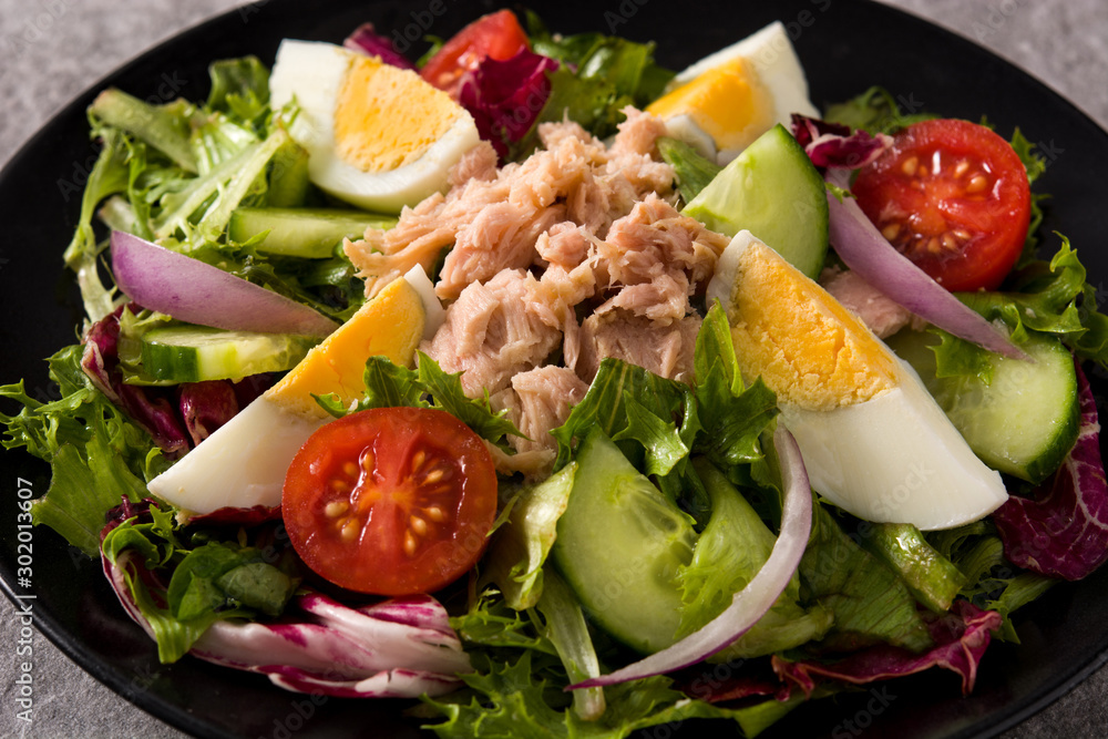 Salad with tuna, egg and vegetables on black plate and gray background. Close up Salad with tuna, egg and vegetables on black plate and gray background. Close up